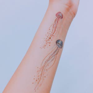 A close up image of two jellyfish tattoos applied across the width of the inner forearm area. One is blue, the other pink and purple. Some areas of metallic gold have caught onto the light, creating a shimmering effect.