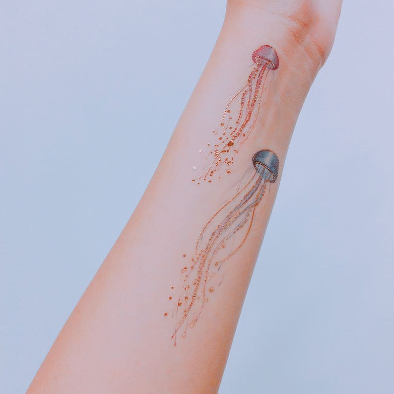 A close up image of two jellyfish tattoos applied across the width of the inner forearm area. One is blue, the other pink and purple. Some areas of metallic gold have caught onto the light, creating a shimmering effect.