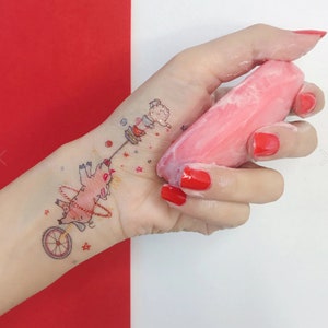 May include: A hand with red nail polish holds a pink bar of soap. A temporary tattoo on the wrist features a pig riding a unicycle and a small figure holding a stack of plates.