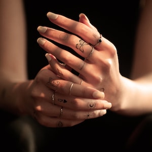 May include: Close-up of hands with multiple small temporary tattoos, including a butterfly, a heart, a diamond, a circle, a crescent moon, a star, a queen of hearts playing card, and an eye. The hands are wearing several thin silver rings.
