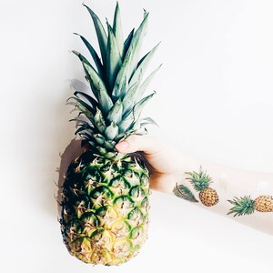 May include: A person is holding a green pineapple with a yellow core. The person has a tattoo of pineapples on their arm.