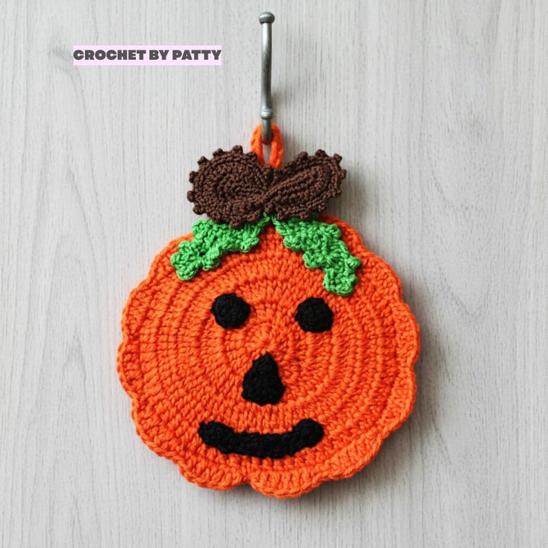 Puede incluir: Un agarrador de crochet naranja con forma de calabaza y cara negra bordada que incluye dos ojos circulares, una nariz triangular y una boca curva. Tiene hojas verdes y un tallo marr&oacute;n en la parte superior. Se lee el texto CROCHET BY PATTY.