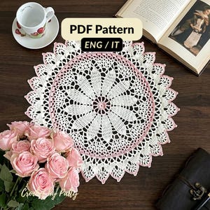 Può includere: Un centrino rotondo all'uncinetto bianco e rosa con un motivo floreale centrale e un bordo smerlato. L'immagine include il testo PDF Pattern ENG / IT. Il centrino è mostrato insieme a una tazza e un piattino in ceramica bianca, un libro e un bouquet di rose rosa.