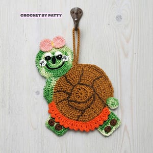 May include: A crochet turtle wall hanging featuring the text CROCHET BY PATTY. The turtle has a green head with a pink bow, a brown shell with dark brown stitching, and an orange ruffled border. It hangs from a brown loop on a metal hook.