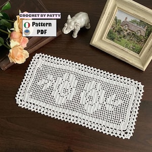 Può includere: Un centrino rettangolare bianco all'uncinetto con un elaborato motivo traforato. È posizionato su una superficie di legno scuro, con una piccola statuina di elefante in ceramica e un'immagine incorniciata sullo sfondo. Il testo "CROCHET BY PATTY" e "Pattern PDF" sono visibili.