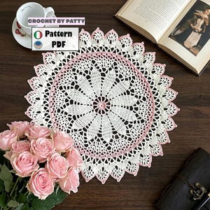 Può includere: Un centrino all'uncinetto bianco e rosa fatto a mano con un motivo floreale. Il centrino è circolare con un bordo smerlato e un bordo rosa. Il testo "CROCHET BY PATTY" e "Pattern PDF" sono visibili.