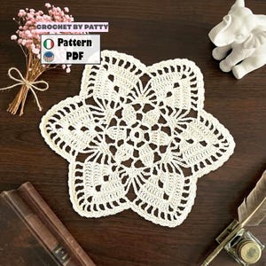 May include: Cream-colored, star-shaped crochet doily with intricate openwork design. The doily is handmade and sits on a dark wooden surface. The image also includes a small bouquet of pink flowers and the text "CROCHET BY PATTY".