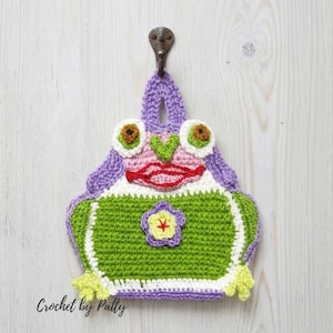 Könnte beinhalten: Ein gehäkelter Topflappen in Froschform mit einer violetten Aufhängeschlaufe, einem weiß-grünen Körper, roten Lippen und einer gelben Blütenapplikation in der Mitte. Der Text Crochet by Patty ist unten links schwarz aufgedruckt.