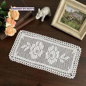 May include: A white, rectangular crocheted doily with a decorative border. The doily is displayed on a dark wooden surface, alongside a small elephant figurine, a framed picture, and a bouquet of roses. The text "CROCHET BY PATTY" is visible.