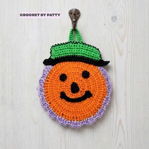 Può includere: Una presina rotonda all'uncinetto a forma di zucca con un cappello nero e verde, caratterizzata da un volto nero ricamato con occhi, naso e sorriso. La zucca è bordata da un bordo smerlato color lilla e include un occhiello. Testo: CROCHET BY PATTY.
