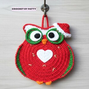 Op de afbeelding: Een rode gehaakte uil pannenlap met een wit hart op de borst, groen omrande ogen met zwarte pupillen, een oranje snavel en pootjes, en een rode kerstmuts met een witte rand. De tekst CROCHET BY PATTY is bovenaan in de afbeelding zichtbaar.