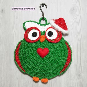 Può includere: Una decorazione a forma di gufo all'uncinetto verde con un cuore rosso sul petto, becco e zampe arancioni e grandi occhi con bordi rossi. Indossa un cappello da Babbo Natale rosso e bianco ed è appeso a un gancio di metallo nero. Il testo CROCHET BY PATTY è visibile in alto.