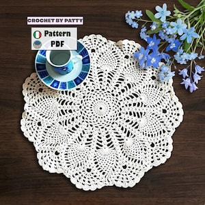 May include: A round white crocheted doily with an intricate pineapple-inspired pattern. A small blue and white ceramic cup sits on the doily, accompanied by a cluster of blue forget-me-not flowers. Text reads CROCHET BY PATTY Pattern PDF.