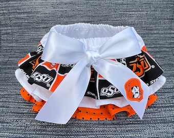 May include: A white ruffled skirt with a black and orange print featuring the letters "OSU". The skirt has a white satin bow tied in the front.