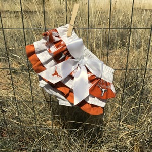 May include: Orange and white ruffled baby bloomers with a white satin bow. The bloomers have a Texas Longhorns print with the team logo.