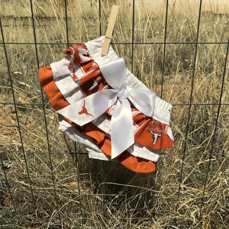May include: Orange and white ruffled baby bloomers with a white satin bow. The bloomers have a Texas Longhorns print with the team logo.