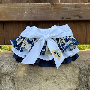 May include: A layered pet skirt featuring a white waistband and a large white bow. The skirt has navy blue and gold fabric with the words "Georgia Tech" and a yellow jacket emblem. The skirt is designed for a pet.