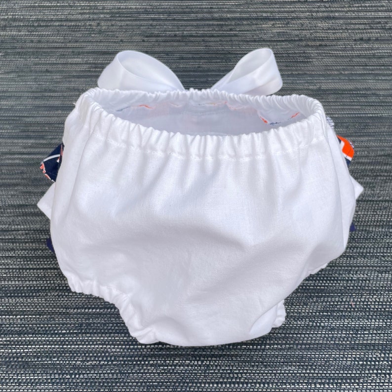 May include: White cotton baby bloomers with a white bow on the back. The bloomers have a ruffled edge and are made of soft, breathable fabric.