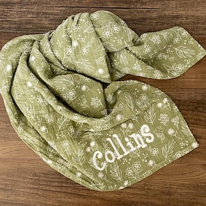 May include: A green muslin swaddle blanket with a white floral pattern and the name "Collins" embroidered in white.