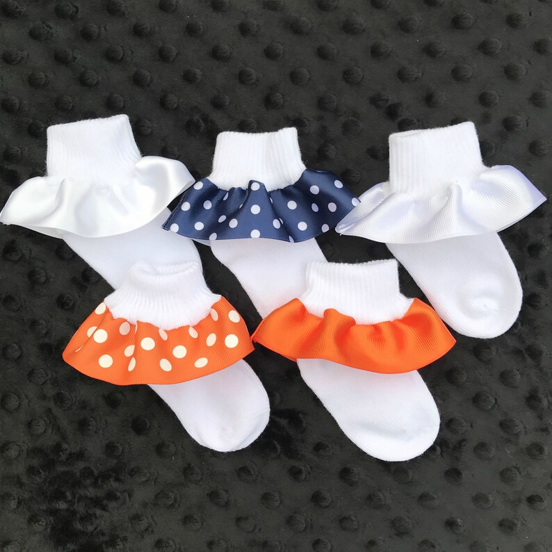 May include: Four pairs of white ankle socks with ruffled edges. The ruffles are made of satin ribbon with polka dots in navy blue, orange, and white.