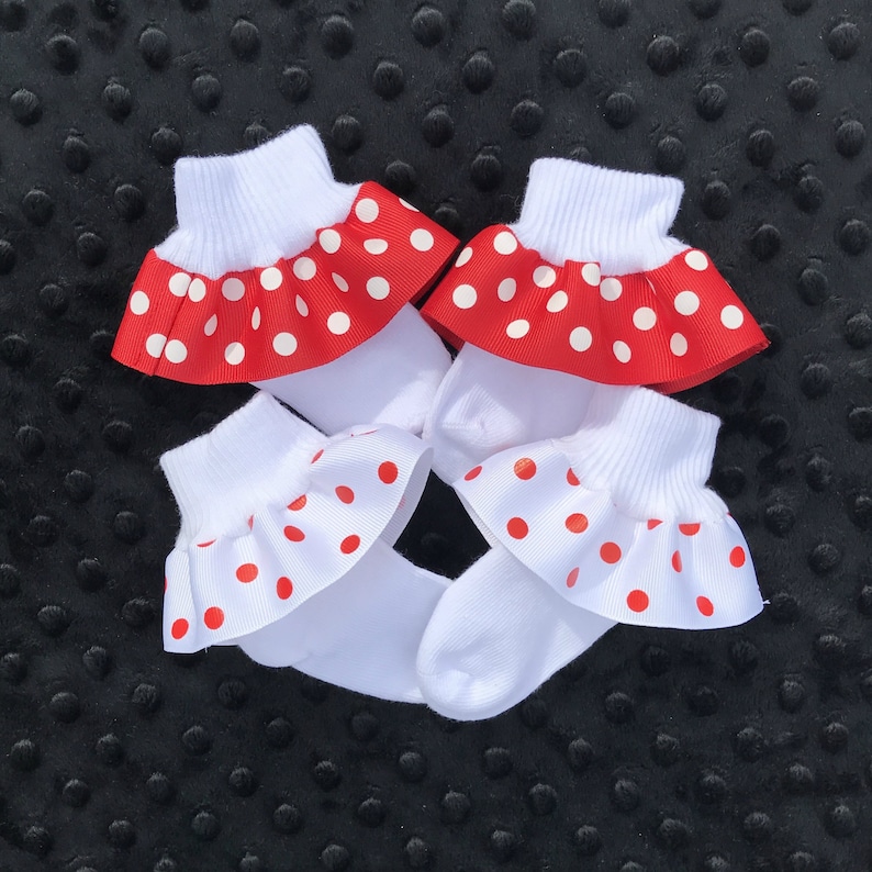 K&ouml;nnte beinhalten: Zwei Paar wei&szlig;e Babysocken mit roten und wei&szlig;en Tupfen-R&uuml;schen am oberen Rand.