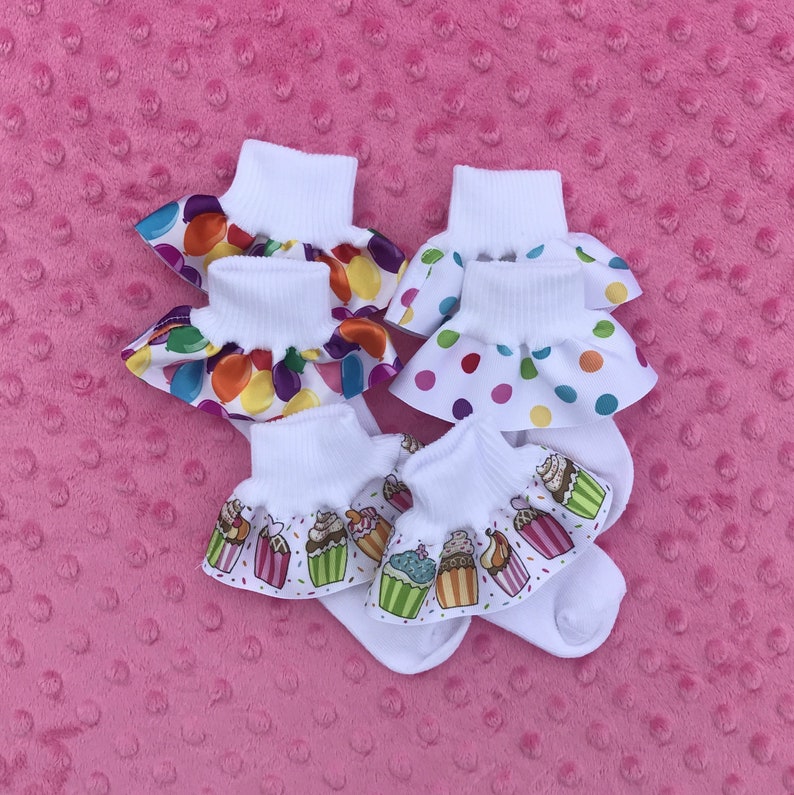 May include: Four pairs of white baby socks with colorful ruffles. The ruffles have different patterns, including polka dots, jelly beans, and cupcakes.