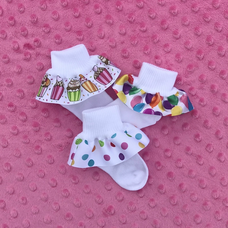 May include: Three pairs of white socks with colorful ruffles. The ruffles have different patterns: cupcakes, confetti, and balloons.