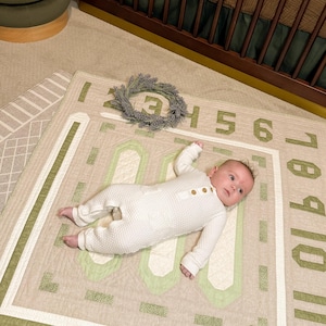 May include: A baby wearing a white waffle knit onesie lies on a green and beige play mat with numbers 1-10 printed on the border. A wreath of lavender is on the mat.