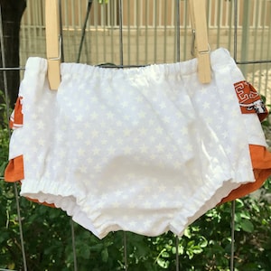 May include: White baby bloomers with a white star pattern. The bloomers are hanging on a clothesline with a brown wooden clothespin. The bloomers are layered over a brown and orange fabric with a white star.