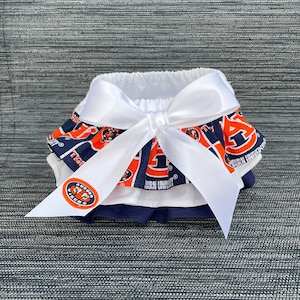 May include: A white ruffled baby bloomers with a white satin bow. The bloomers are decorated with a blue and orange print featuring the Auburn University logo and the words "Auburn Tigers".