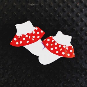 K&ouml;nnte beinhalten: Zwei wei&szlig;e Babysocken mit roten und wei&szlig;en Tupfen-R&uuml;schen. Die Socken liegen auf einem schwarzen Hintergrund.