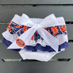 May include: White ruffled diaper cover with a blue and orange Auburn Tigers print and a white satin bow. The diaper cover has a white elastic waistband and a white ruffle trim.