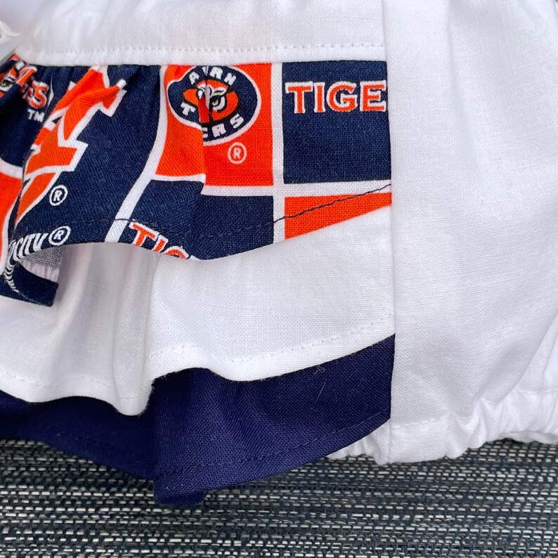May include: A white fabric skirt with a blue and orange ruffle trim. The ruffle has a pattern of a tiger head with the words "Auburn Tigers" and "TIGER" on it.