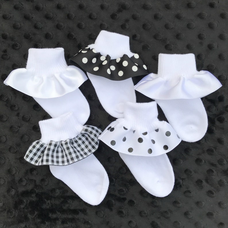 May include: Five pairs of white ankle socks with different black and white patterned ruffles. The ruffles are made of fabric with polka dots, gingham, and solid black.