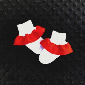 K&ouml;nnte beinhalten: Ein Paar wei&szlig;e Babysocken mit rotem R&uuml;schenbesatz. Die Socken liegen auf einem schwarzen, strukturierten Hintergrund.