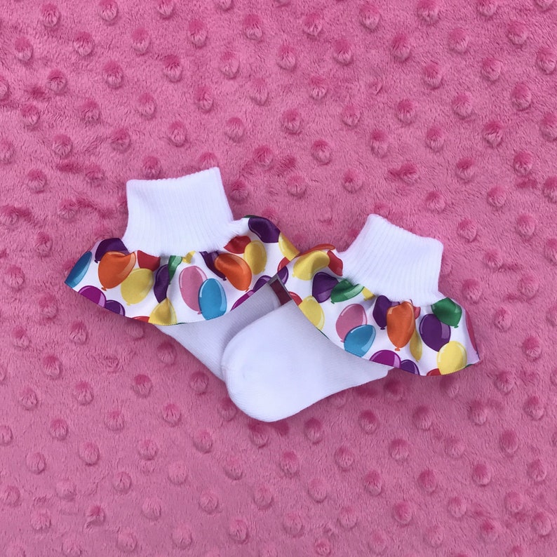 May include: A pair of white ankle socks with a colorful balloon print ruffle trim. The socks are on a pink fuzzy surface.