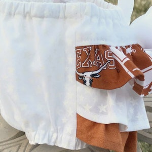 May include: White baby bloomers with a brown and white star pattern. The bloomers have a brown ruffle with the word "TEXAS" and a longhorn steer design.