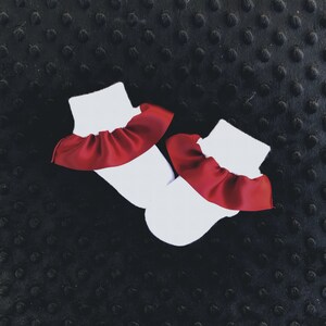 K&ouml;nnte beinhalten: Ein Paar wei&szlig;e Babysocken mit rotem R&uuml;schenbesatz. Die Socken liegen auf einem schwarzen, strukturierten Hintergrund.