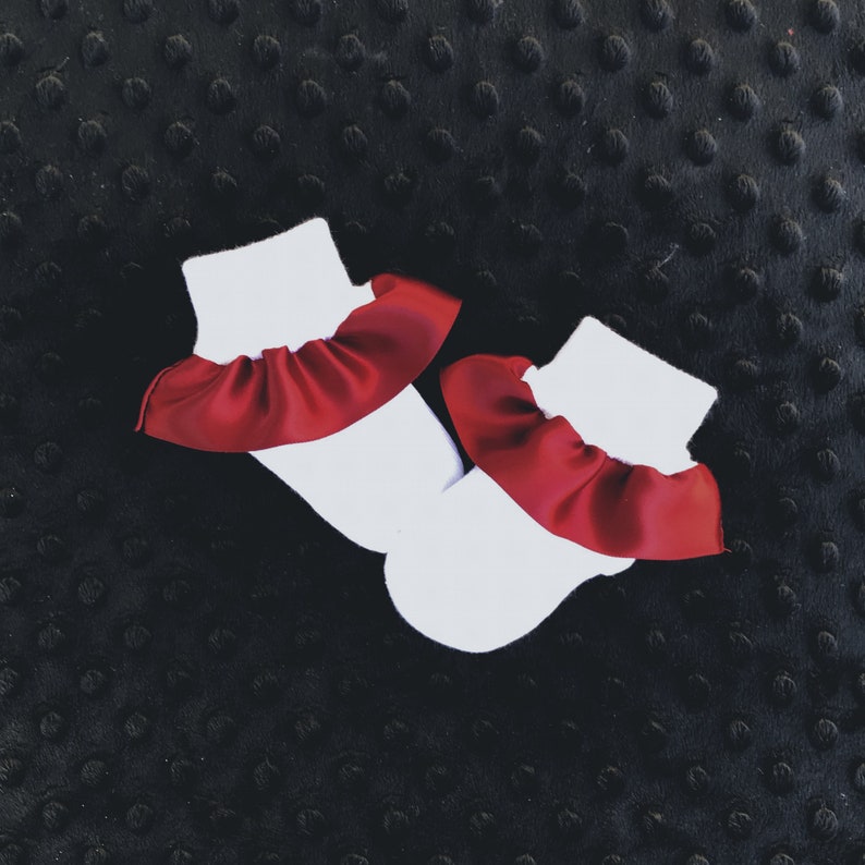 K&ouml;nnte beinhalten: Ein Paar wei&szlig;e Babysocken mit rotem R&uuml;schenbesatz. Die Socken liegen auf einem schwarzen, strukturierten Hintergrund.
