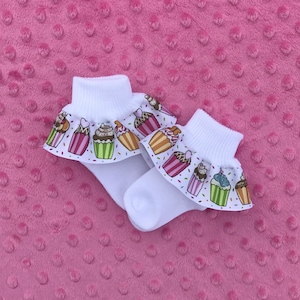 May include: A pair of white baby socks with a cupcake print and a ruffled trim. The socks are on a pink background.
