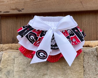 May include: A white, red, and black ruffled skirt with a white bow and a University of Georgia logo. The skirt has a red and white polka dot pattern on the bottom layer.