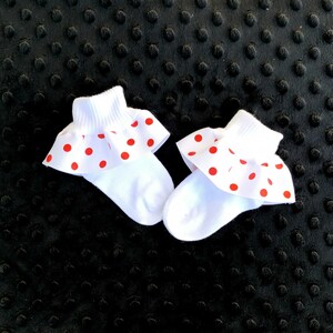 K&ouml;nnte beinhalten: Ein Paar wei&szlig;e Babysocken mit roten Punkten und einem R&uuml;schenbesatz. Die Socken liegen auf einer schwarzen, strukturierten Oberfl&auml;che.