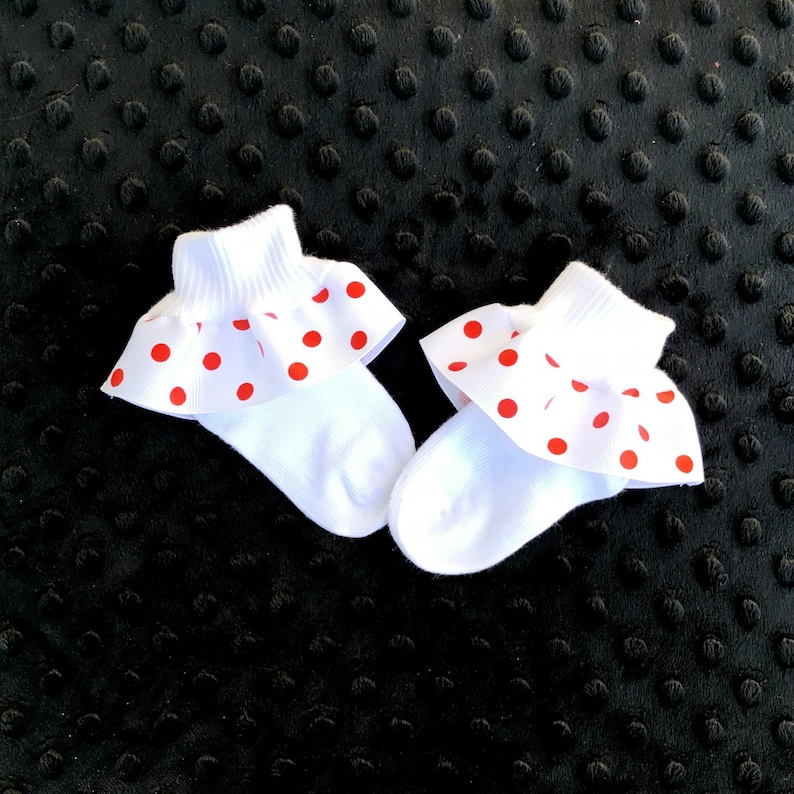 K&ouml;nnte beinhalten: Ein Paar wei&szlig;e Babysocken mit roten Punkten und einem R&uuml;schenbesatz. Die Socken liegen auf einer schwarzen, strukturierten Oberfl&auml;che.