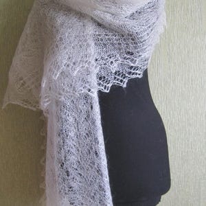 May include: A delicate, white, lacy shawl draped over a black mannequin. The shawl has a soft, airy texture and a scalloped edge. The intricate openwork design is visible throughout the fabric, creating a light and elegant look.