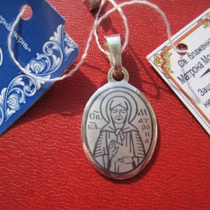May include: Silver pendant featuring a black and white image of Saint Matrona of Moscow. The pendant is oval shaped and has a silver chain attached. The text on the pendant reads "Св. Бл. Матрона".