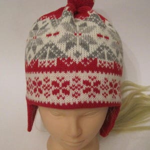 May include: Red and white knitted hat with a gray snowflake pattern and a white pom pom on top. The hat has ear flaps.