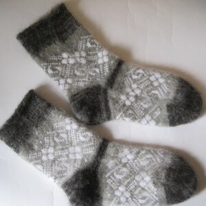 K&ouml;nnte beinhalten: Ein Paar flauschige Strickstr&uuml;mpfe mit einem geometrischen Muster in Grau und Wei&szlig;. Die Socken haben einfarbige, dunkelgraue B&uuml;ndchen, Fersen und Zehenbereiche und eine weiche, strukturierte Optik mit einem sich wiederholenden Rauten- und Blumenmuster.