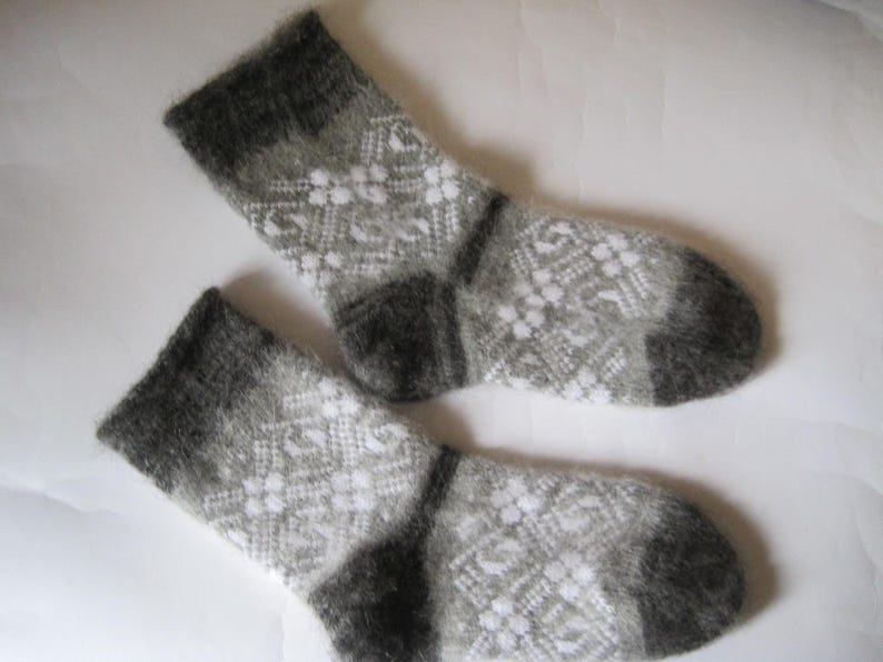 K&ouml;nnte beinhalten: Ein Paar flauschige Strickstr&uuml;mpfe mit einem geometrischen Muster in Grau und Wei&szlig;. Die Socken haben einfarbige, dunkelgraue B&uuml;ndchen, Fersen und Zehenbereiche und eine weiche, strukturierte Optik mit einem sich wiederholenden Rauten- und Blumenmuster.