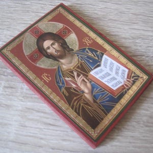 May include: A red wooden icon with a gold frame depicting Jesus Christ holding a book. The book has text in a foreign language. The icon has a cross behind Jesus's head.