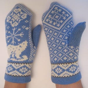 May include: A pair of blue knitted mittens with white and brown geometric and animal patterns. The mittens feature a snowflake and polar bear design on one and a diamond pattern on the other. The cuffs and thumbs are solid blue.